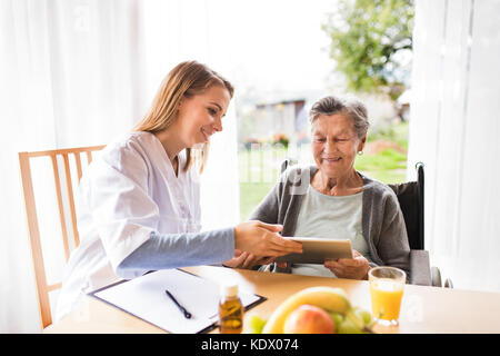 Gesundheitsbesucher und eine Seniorin mit Tablet während Hausbesuch. Eine Krankenschwester, die mit einer älteren Frau spricht. Stockfoto