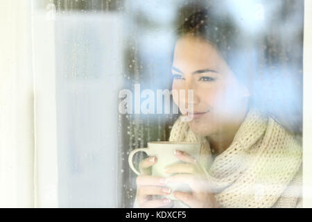 Porträt eines Mädchens, das durch ein Fenster mit einer Tasse Kaffee in einer regnerischen Tag der Winter Stockfoto