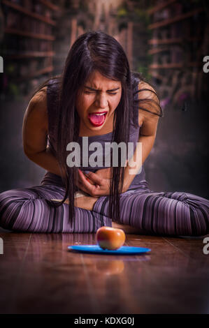 Schöne und einsame Mädchen leiden unter appetitanregend, Schmerzen in seinem Magen wenn sie sieht eine Apple über einen blauen Platte, in einer verschwommenen Hintergrund Stockfoto