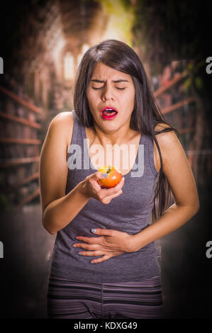 Schöne und einsame Mädchen leiden unter appetitanregend, Schmerzen in seinem Magen wenn Sie sich einen Apfel und Tun ekelhaft Gesichter, in einem verschwommenen Hintergrund Stockfoto