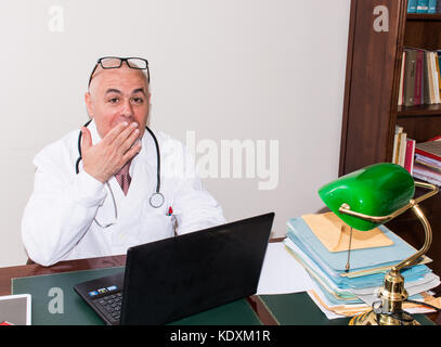 Arzt in seinem Studio am Schreibtisch, am PC, mit Brille. Neue Technologien verwenden. In seiner professionellen Studio, er sitzt an antiken Schreibtisch, und eine grüne Lampe Stockfoto