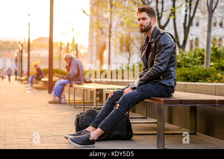 Ein hübscher Junge Hipster Mann sitzen und schaut sich um auf einer Bank in den Sonnenuntergang Stockfoto