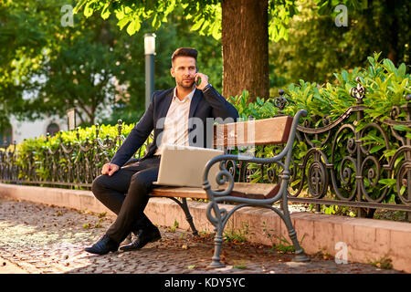 Ein hübscher junger Geschäftsmann Standortwahl auf einer Bank mit seinem Laptop an einem sonnigen Straße neben einem Park, der gerade sein Telefon Stockfoto