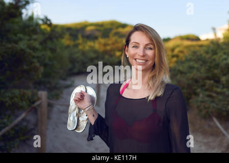 Attraktive schlanke blonde Frau ihre Schuhe tragen als sie Spaziergänge an einem Sandstrand weg Lächeln glücklich an der Seite im Sommerurlaub Stockfoto