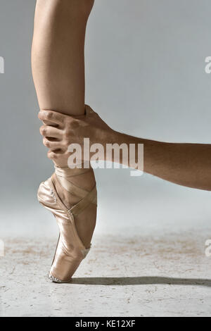 Die Hand des Mannes hält das Bein des Ballerina auf dem grauen Hintergrund im Studio. Mädchen trägt beige Spitzenschuhe und steht auf der Zehe. closeup. Vertikale Stockfoto