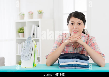 Elegante attraktive Hausfrau auf dem Bügelbrett an Kamera lächeln, wenn Sie alle allgemeine Ordnung und Sauberkeit zu Hause fertig Suchen lehnend. Stockfoto