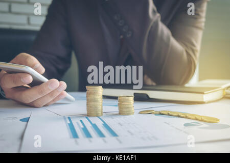 Geschäftsmann mit smart phone mit gestapelten Münzen im Büro Schreibtisch und vielen Dokument Datengraphik auf Morgen angeordnet, das Konzept. Stockfoto
