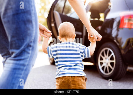 Nicht erkennbare junge Mutter helfen Ihr kleines Baby Boy gehen. Erste Schritte, in der Nähe ein Auto. Sicht nach hinten. Stockfoto