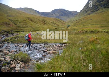 Einsame männliche Walker Überquerung des Flusses allt na muidhe in gleann - leac-na-muidhe mit der munro sgor na h-ulaidh in der Ferne in den schottischen Highlands. Stockfoto