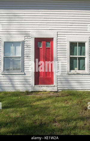 Eine helle rote Tür auf einem alten weiß bemalten Schindeln Holz gerahmt Farm House. Stockfoto