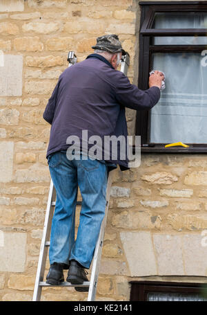 Rüstige alte Mann mit Hut, Jacke und Jeans, auf eine Leiter, ein Fenster im Obergeschoss eines Hauses aus Stein. Langtoft, England, UK. Stockfoto