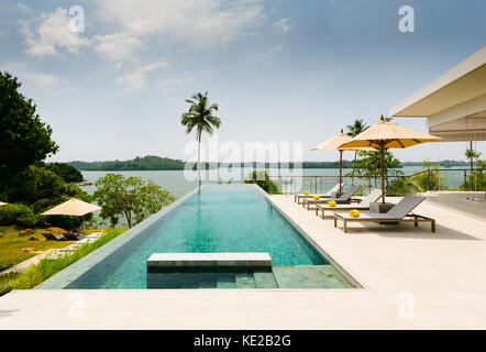 Infinity Pool mit Blick auf einen See, der von Palmen, Liegestühlen und Sonnenschirm grenzt, in der modernen Boutique Hotel Resort, Tangalle, Sri Lanka Stockfoto