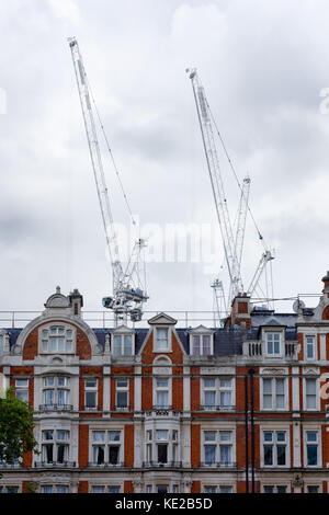 Zwei weiße Krane entstehen gegen einen bewölkten Himmel und hinter einer Reihe von typischen Backsteinhäuser in London, UK. Stockfoto