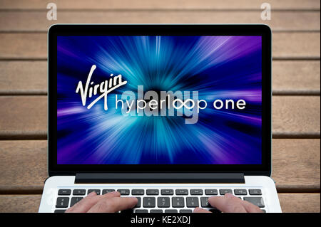 Die Jungfrau Hyperloop ein Logo wird auf dem Bildschirm des MacBook Pro Laptop gezeigt, Schuß gegen eine Holzbank einschließlich der Finger des Mannes (nur redaktionell). Stockfoto