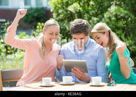 Gruppe von Aufgeregt Freunde auf der Suche nach digitalen Tablet mit Tasse Kaffee auf Tisch Stockfoto