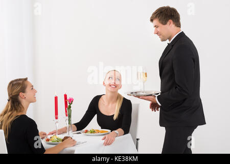 Gerne Kellner serviert Glas Champagner zu weiblichen Freunde im Restaurant mit Essen auf dem Tisch Stockfoto