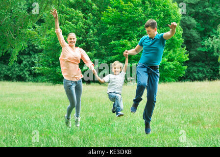 Glückliche Eltern halten einander die Hände Springen mit Ihrem Sohn Im Park begeistert Stockfoto