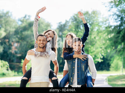 Zwei Paare von Menschen in den Park, Mädchen sind wellenartig. Lustig junge Frauen sitzen auf dem Rücken der Jungs, Freuen und fröhlich begrüßen. glückliche Liebhaber pi Stockfoto