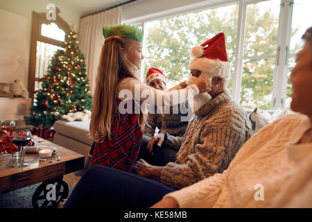 Kleines Mädchen, fake Weihnachtsmann Bart und Hut auf ihr Großvater. Familie sitzen zusammen im Wohnzimmer am Heiligabend. Stockfoto