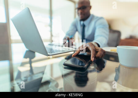 Mann bei der Arbeit am Laptop an seinem Schreibtisch im Büro zu sitzen. Der Mensch erreicht sein Handy auf den Tisch gelegt. Stockfoto