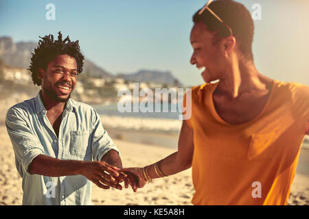 Verspieltes junges Paar, das die Hände am sonnigen Sommerstrand hält Stockfoto