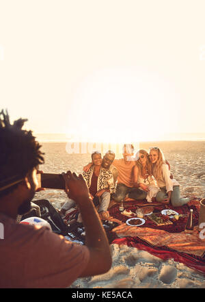 Junger Mann mit Kameratelefon, der Freunde beim Picknick fotografiert Sonniger Sommerstrand Stockfoto