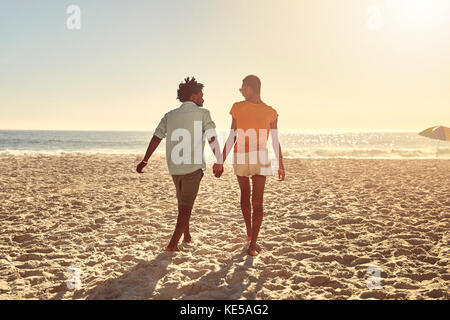 Junges Paar, das Hände hält und am sonnigen Sommerstrand spazierengeht Stockfoto