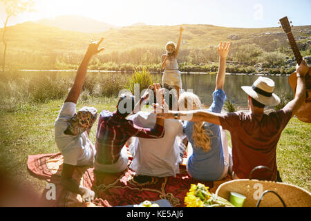 Frau mit Kameratelefon fotografiert Freunde mit erhobenen Armen Sonniger Sommer am Flussufer Stockfoto