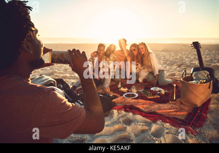 Junger Mann mit Kameratelefon, der Freunde beim Picknick fotografiert Sonniger Sommerstrand Stockfoto