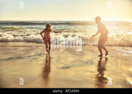 Verspieltes junges Paar, das im sonnigen Sommer im nassen Sand läuft meeresstrand Stockfoto
