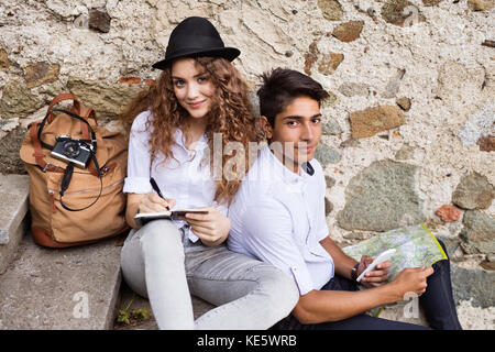 Zwei schöne junge Touristen mit Smartphone, Karte und Kamera in der Altstadt. Teenager sitzen auf Treppen, schreib.. Stockfoto