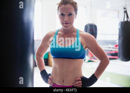 Portrait selbstbewusste, robuste Boxerin, die im Fitnessstudio beim Boxsack steht Stockfoto