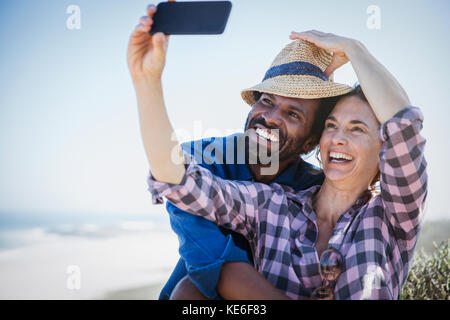 Verspieltes, lächelndes multiethnisches Paar, das Selfie mit dem Kameratelefon am sonnigen Sommerstrand macht Stockfoto