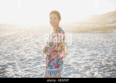 Portrait lächelnde, selbstbewusste Brünette Frau in Badeanzug bedeckt auf sonnigen Sommer Sonnenuntergang Strand Stockfoto