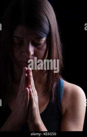 Gläubigen athletische Frau beten, Hände in der Anbetung zu Gott gefaltet, Kopf und Augen in religiöser Inbrunst geschlossen. schwarzer Hintergrund. Gebet portrait Stockfoto