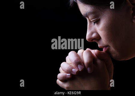 Treue Frau zu beten, die Hände gefaltet in der Anbetung mit Kopf nach unten zu Gott und Augen in religiöser Inbrunst geschlossen, auf einem schwarzen Hintergrund/Gebet portrait Stockfoto