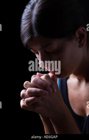 Low Key Porträt einer gläubigen Frau zu beten, die Hände gefaltet in Gottesdienst, Kopf und Augen in religiöser Inbrunst geschlossen, auf einem schwarzen Hintergrund. Stockfoto