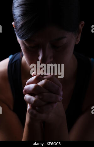 Low Key Porträt einer gläubigen Frau zu beten, die Hände gefaltet in Gottesdienst, Kopf und Augen in religiöser Inbrunst geschlossen, auf einem schwarzen Hintergrund. Stockfoto