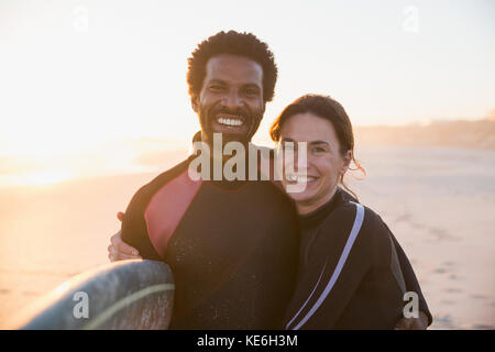 Portrait lächelndes, selbstbewusstes multiethnisches Paar mit Surfbrett am sonnigen Strand bei Sonnenuntergang Stockfoto