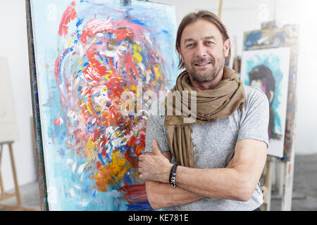 Portrait lächelnder, selbstbewusster Künstler, der bei abstrakter Malerei im Atelier der Kunstklasse steht Stockfoto