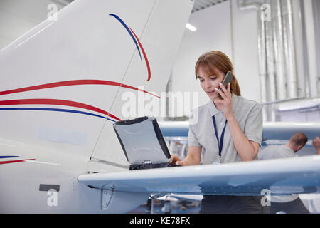 Weibliche Flugzeugingenieurin, die am Laptop arbeitet und über Zelle spricht Telefon im Hangar Stockfoto