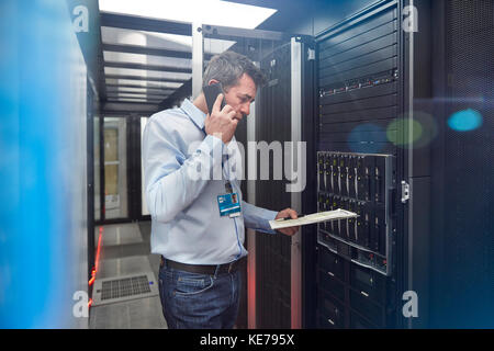 Männlicher IT-Techniker mit Clipboard im Gespräch auf Handy bei Panel im Serverraum Stockfoto