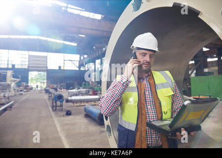 Männlicher Ingenieur mit Laptop, der auf dem Handy in Stahl spricht Werk Stockfoto