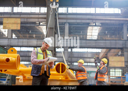 Männlicher Vorarbeiter mit Klemmbrett, der in der Nähe der Arbeiter in der Fabrik steht Stockfoto