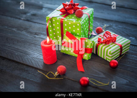 Fröhliches Bild mit einem Bündel grün eingepackte Geschenke, mit roten Bändern und Schleifen gebunden, durch Weihnachtsverzierungen umgeben und eine brennende Kerze, auf einer hölzernen ta Stockfoto