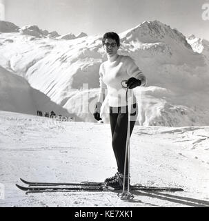 1960, historische Bild eines attraktiven jungen weiblichen Skifahrer, leicht bekleideten, mit Ski, Handschuhe und Skistöcke, stehend auf einer Piste am Bardbou mit den Bergen hinter ihr, Frankreich. Stockfoto