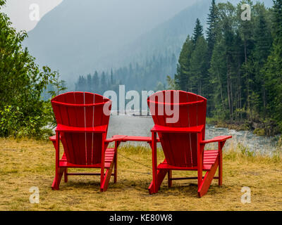 Zwei rote Adirondack Stühle sitzen auf der Wiese mit Blick auf den Columbia River und Wald bedeckte Berge; Revelstoke, British Columbia, Kanada Stockfoto