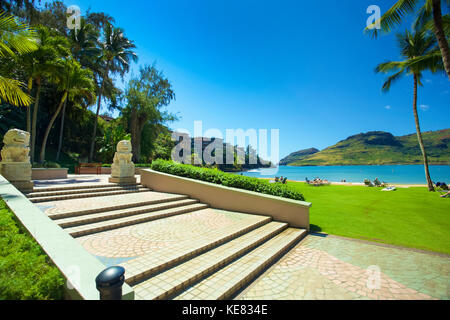 Touristen sitzen auf einem Strand mit einer Hawaiian Resort entlang einer Bucht mit hellen grünen Gras, blauer Himmel und Palmen. Stockfoto