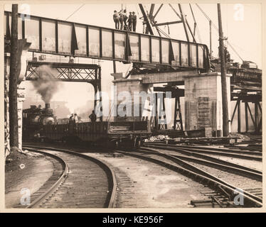 Dieses Foto zeigt Arbeiter, die auf einem Gestell stehen, während ein Zug darunter fährt. Das Bild fängt industrielle Arbeit und Transport ein, mit einem Fokus auf die Eisenbahn und ihre Arbeiter während des frühen 20. Jahrhunderts. Stockfoto