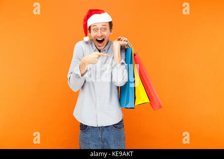 Positive glück Mann an der Kamera und toothy Lächeln suchen. schockiert Mann mit vielen Taschen und zeigen Sie mit Fingern auf Sie. Studio orange Wand geschossen. Stockfoto